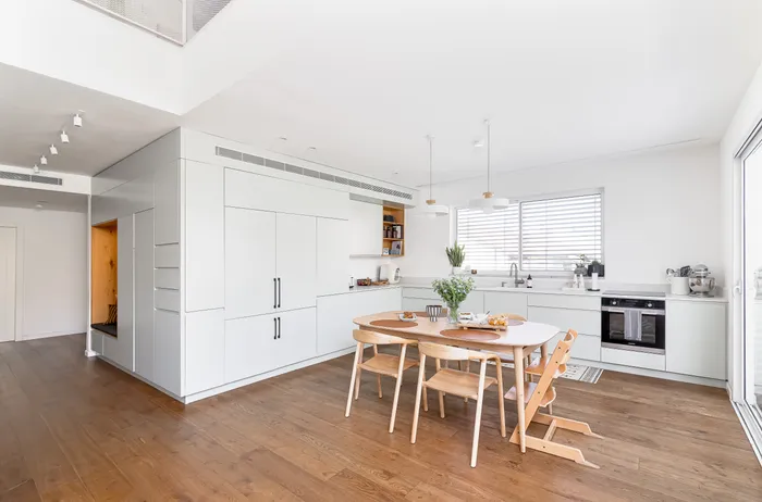 מטבח לבן עם ארונות גבוהים, שולחן אוכל מעץ ונישת אלון מוארת